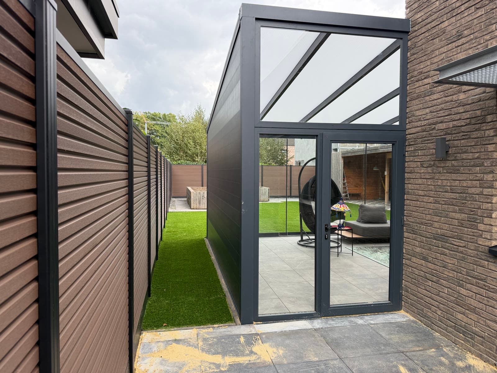 Smalle aanbouw met glazen dubbele deur langs de schutting, naast de tuinkamer